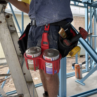 Person holding two cans of Oatey Primer in a red Constricta Multi Pot Grip on a construction site.